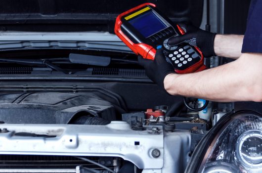 Closeup mechanic hands are holding special diagnostic equipment, scanner at vehicle with open hood. Repairman is repairing car and detecting problems at service station car repair shop.